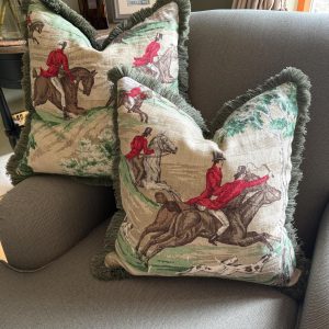 Two Tally Ho Huntmaster Collection cushions with green ruched fringe displayed on a grey armchair. The cushions feature a vintage-style fox hunting scene with riders in red coats on horseback, adding a classic equestrian charm to the elegant living room setting.