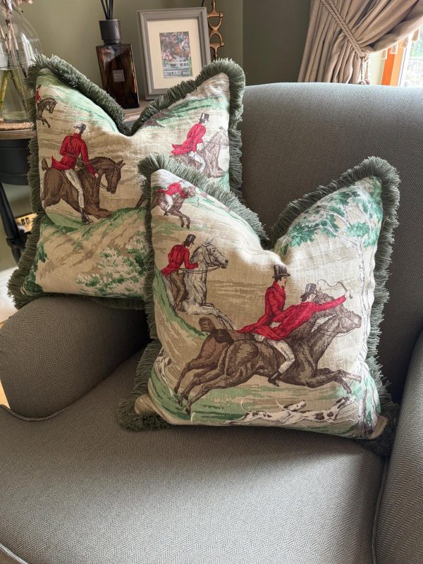 Two Tally Ho Huntmaster Collection cushions with green ruched fringe displayed on a grey armchair. The cushions feature a vintage-style fox hunting scene with riders in red coats on horseback, adding a classic equestrian charm to the elegant living room setting.